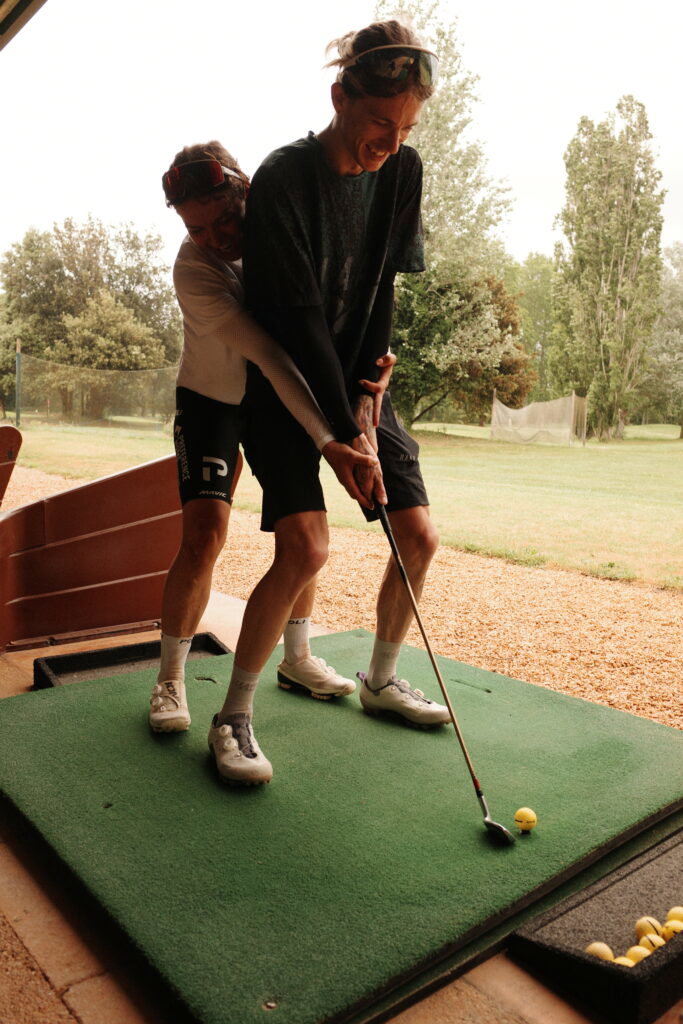 Tailfin cyclists in cycling gear laughing while playing golf at a driving range.